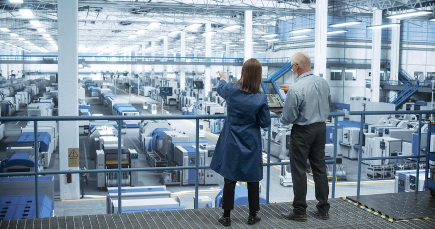 Male and Female Engineers Standing with Their Back to Camera, Using Laptop Computer and Talking in a Factory Facility with Equipment Producing Modern Electronic Components for Different Industries