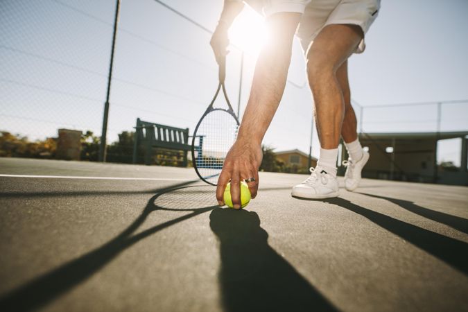 Ground level view of a man picking a tennis ball up on a  sunny day