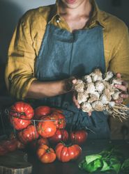 Woman in yellow holding garlic, with wire basket of large tomatoes
