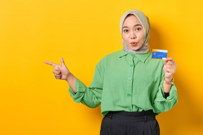Shocked Muslim woman in headscarf and green blouse holding credit card and pointing aside