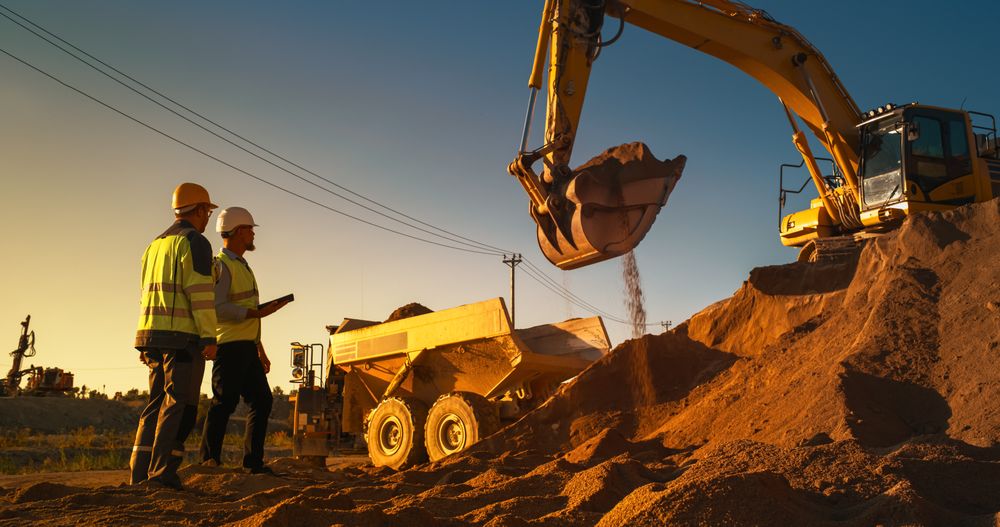 Real Estate Investor With Tablet Computer And Project Manager Talking On Construction Site Of Apartment Building. Colleagues Wearing Hard Hats And Watching Heavy Machinery Working. Sunny Day