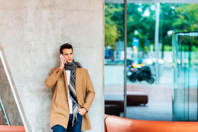 Serious man leaning on wall outside speaking on phone and wearing a camel coat 