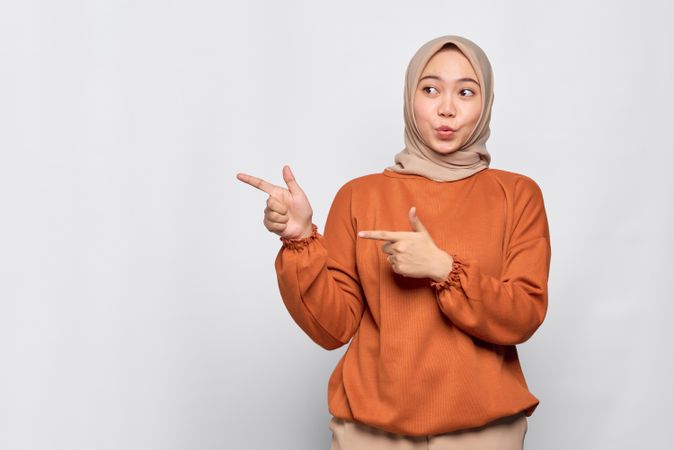 Surprised Muslim woman in headscarf and orange sweater pointing finger to blank copy space