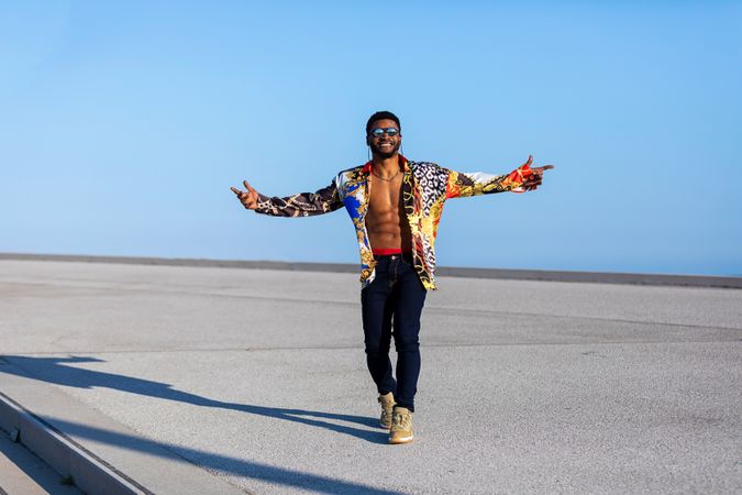 Black man with open arms wearing eyeglasses and colorful shirt against blue sky on sunny day