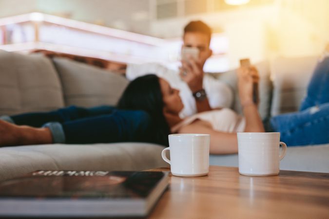 Two coffee cups on table with couple relaxing in background on couch