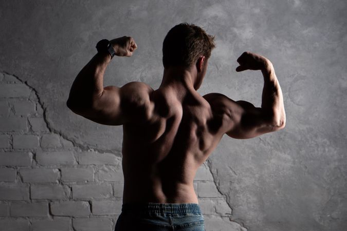 Shirtless man in jeans flexing biceps against textured gray wall.