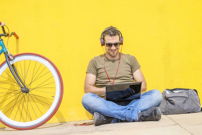 Man with red headphones sitting outside next to bicycle looking at laptop