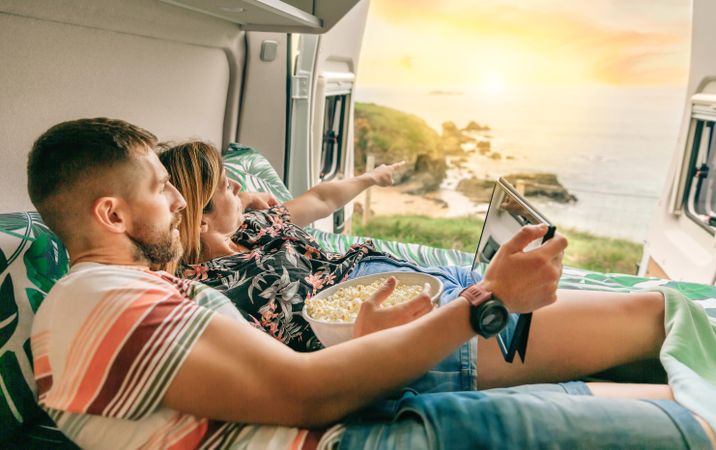 Male and female reclining on bed in motorhome enjoying the view with popcorn and digital tablet
