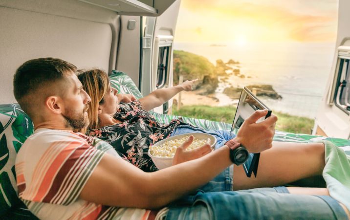 Male and female reclining on bed in motorhome enjoying the view with popcorn and digital tablet