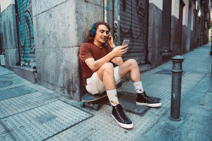 Relaxed man in headphones using smartphone while resting on longboard on street