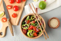 Fresh salad with chopsticks and kitchen accessories on grey background, top view