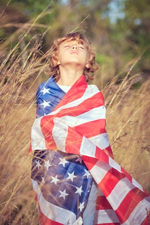Adorable boy wrapped in American flag standing with closed eyes