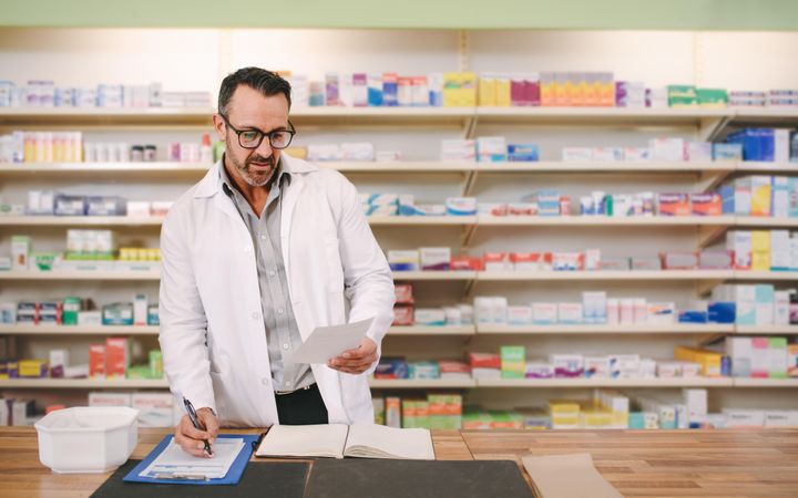 Mature male chemist reading a prescription paper and writing on clipboard at counter