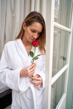 Young woman in white terry bathrobe sniffing rose at window