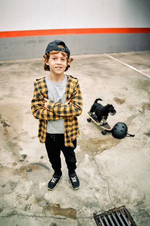Cute boy standing on street near small dog on skateboard