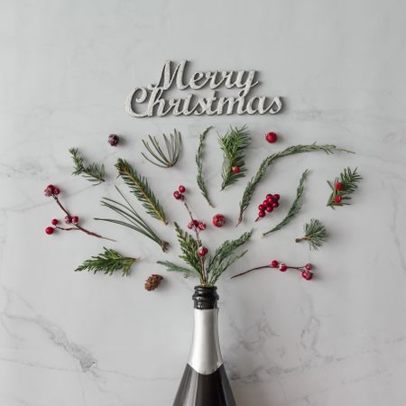 Champagne bottle with winter foliage on marble background with the words “Merry Christmas”