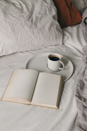 Cup of coffee, espresso. Open book, diary mockup on white bed sheet. Breakfast in bed concept. Blurred muslin, linen pillows, blanket. Scandinavian bedroom. Vertical top view. Lifestyle banner.