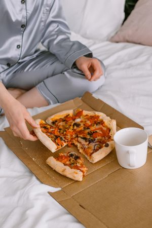 Cropped image of a person in a pajama eating pizza on bed