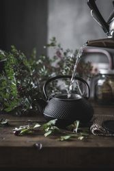 Woman pouring green tea in mug on wooden table with green herb