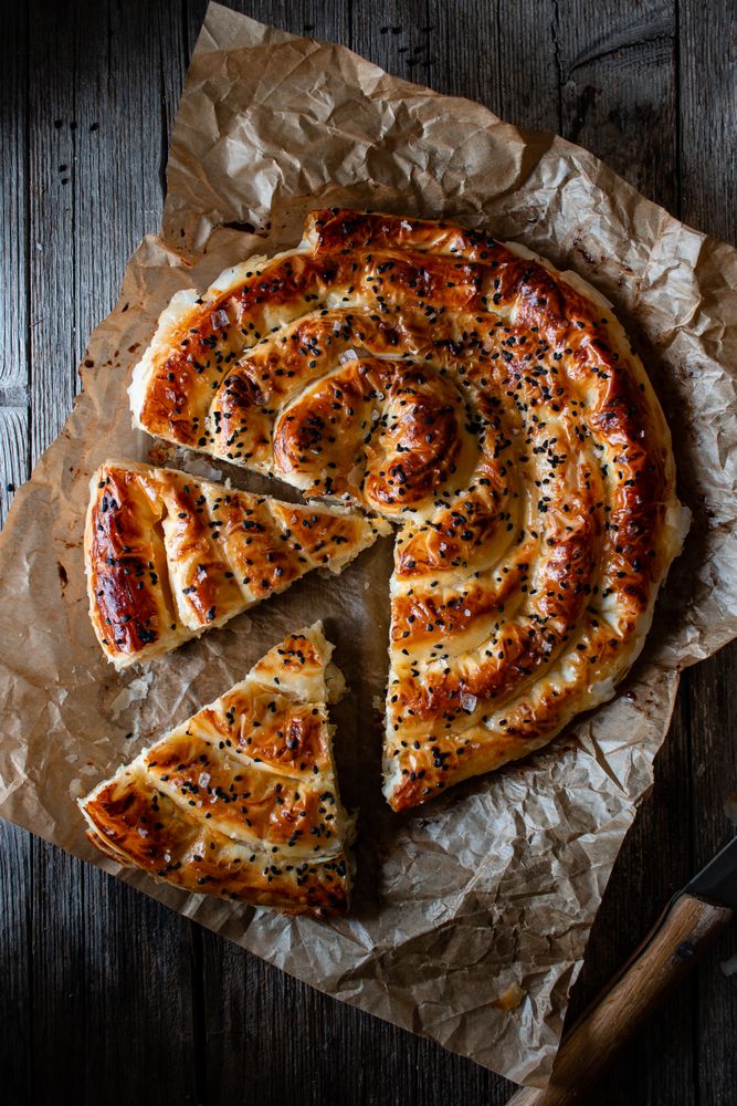 From above crop cook holding hot baking dish with fresh puff pastry and serving piece of borek on plateFaceless person serving homemade borek on wooden table
