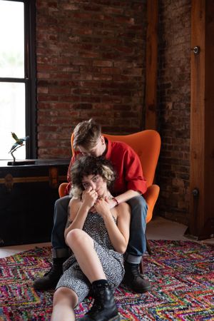 Woman in orange chair kisses her partner’s head while partner looks at camera