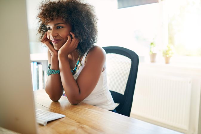 Satisfied curly haired female at her home office desk reading computer screen