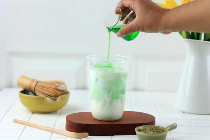 A Hand Pours Green Matcha Syrup into a Layered Green Beverage in a Plastic Cup