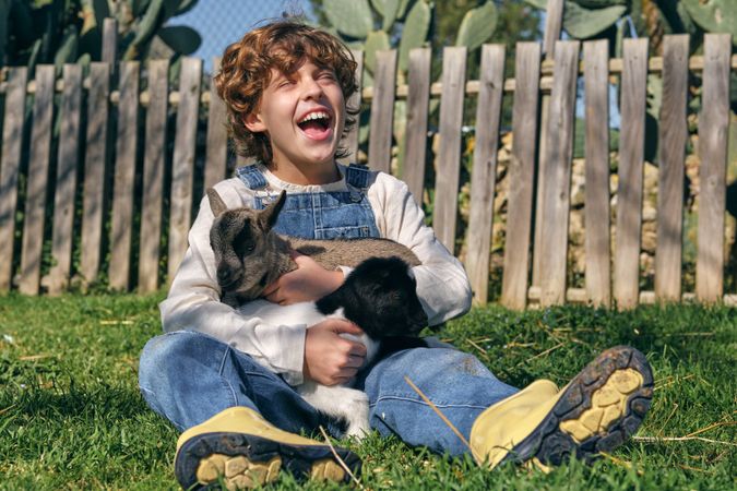 Joyful boy hugging cute baby goats and laughing sitting on lawn