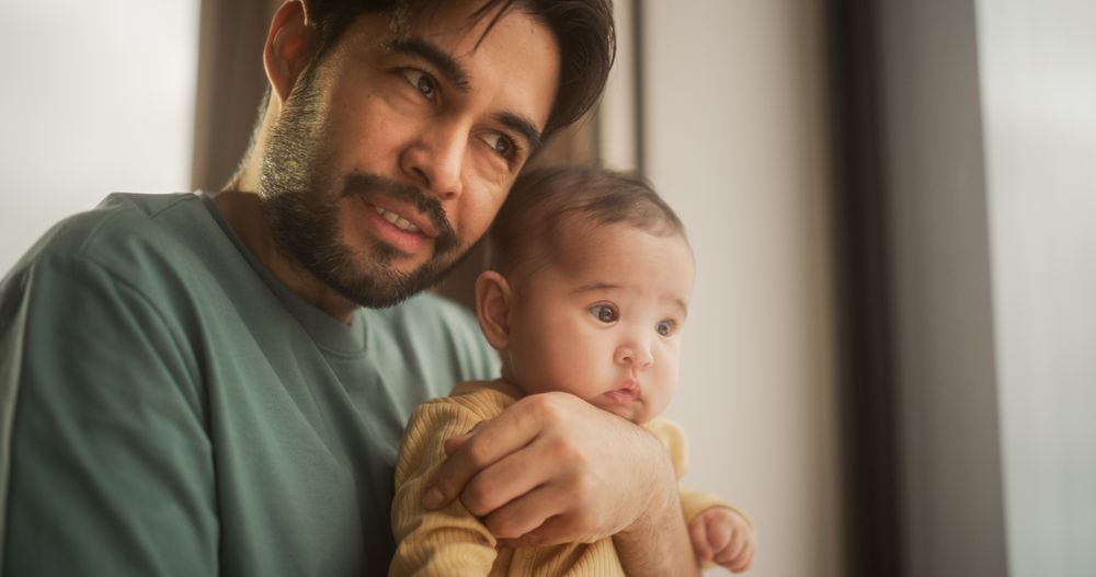 Portrait of Fatherly Love: A Young Father Holding Little Baby in his Arms as He Talks and Watches Through a Window.
