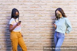 Two girls with facemasks standing apart holding phone beside wall bYrj90