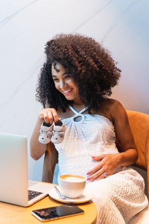 Smiling pregnant Black woman showing baby booties to laptop