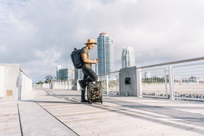 Young man wearing a hat, With A Suitcase and a backpack is searching for directions ion the phone.