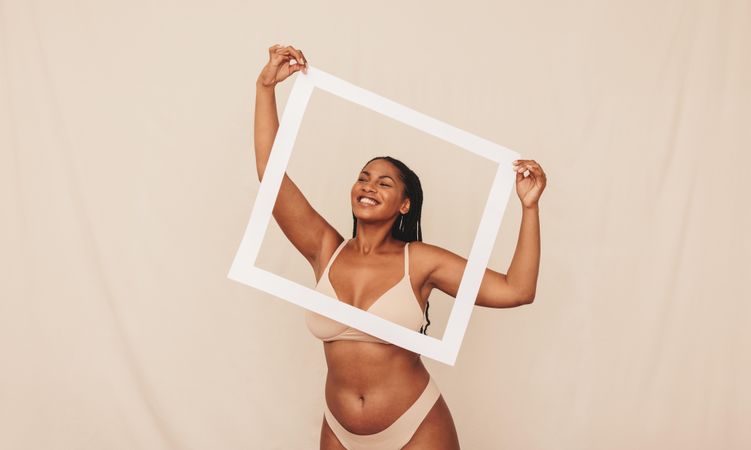 Playful curvy woman wearing beige underwear smiling through a picture frame in a studio