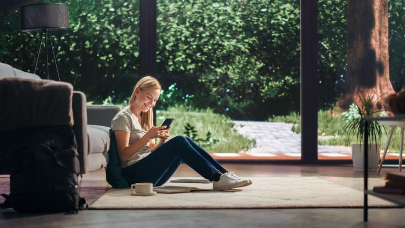 Portrait of Beautiful Young Adult Female with Blond Hair Sitting on a Floor and Using Smartphone in Sunny Living Room.