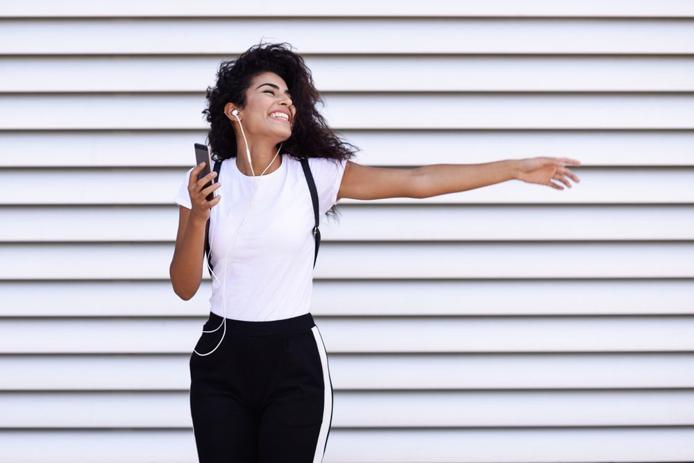 Arab woman in sport clothes with curly hair standing in front of wall dancing with phone