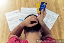 Back view of frustrated man holding credit cards sitting at a table with documents 428Wx0