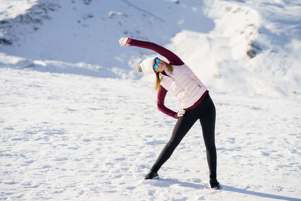 Full body of female in hat, polarized sunglasses, stretching sideways on on snowy mountain
