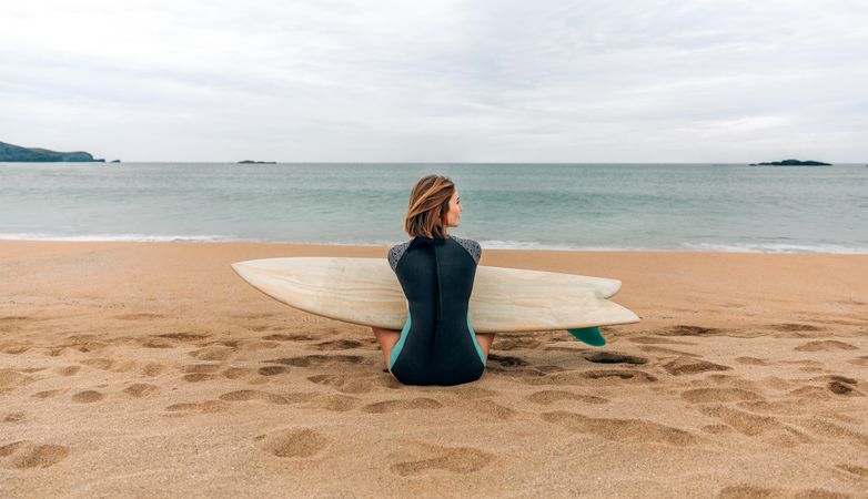 Back of female sitting on the shore with surfboard gazing at the ocean