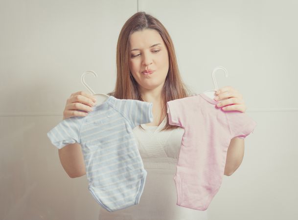 Pregnant woman holding bodysuits  for a girl or a boy in soft pastel colors