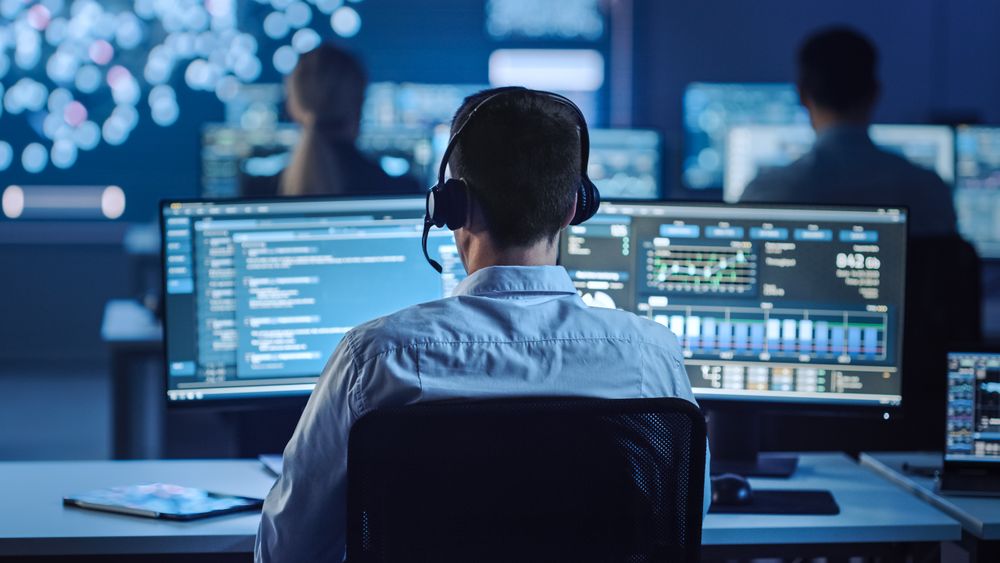 IT Specialist With Headset Monitors Real-time Data on Multiple Screens in Blue-lit Operations Center. Colleagues Work in Background Amid Displays Showing Complex Information.