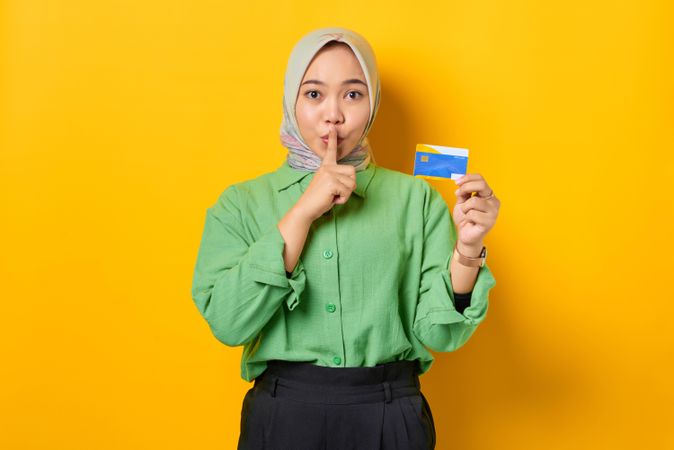 Smiling Muslim woman in headscarf and green blouse saying “shhh” and holding her credit card
