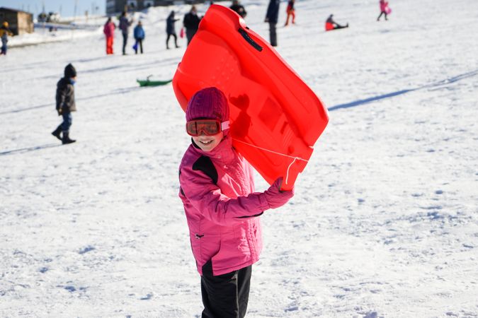 Child in pink snow suit holding sled at busy resort