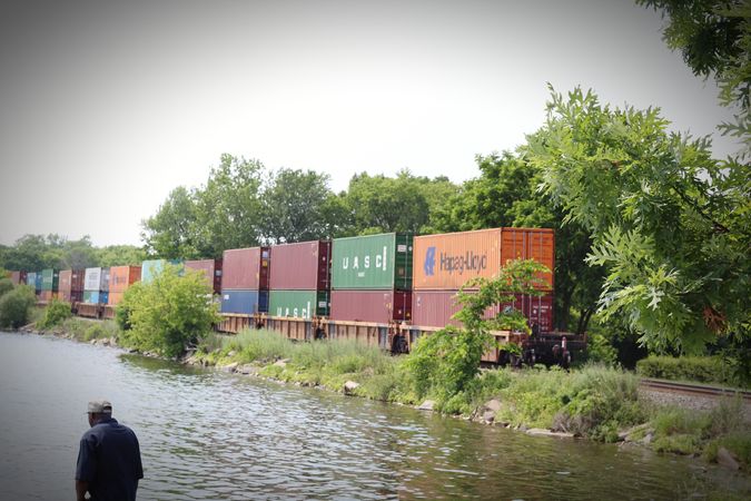 Back view of man standing on shoreline of a river near cargo container on train rail