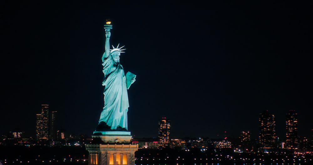 Panoramic Aerial Image of an American Symbol of Freedom. Cinematic Helicopter Pass by the Statue of Liberty at Night. Landmark New York Monument with Manhattan Skyscrapers in the Background