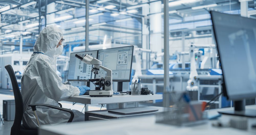 Modern Factory: Industrial Engineer Working on Desktop Computer in Clean Sterile Coveralls. Scientist Using a Microscope, Developing Advanced Solutions for High Tech Medical Vaccine and Gene Research