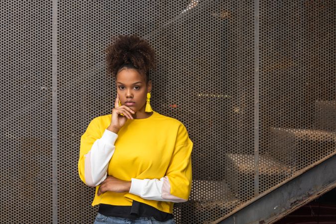 Woman with curly hair wearing yellow long sleeve shirt standing beside gray metal fence