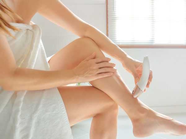 Young woman depilates her leg with an electric epilator device in the bathroom.
