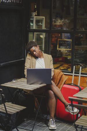 Black woman talking on smartphone and sitting at table with laptop in cafe