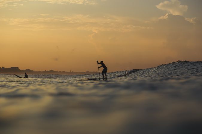 Men with paddle on surf board between waving water at sunset