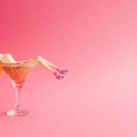 Doll bathing in martini glass full of gold glitter on pink background